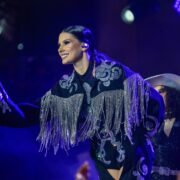 Ana Castela, que tem 20 anos de idade, está com roupa country preta com franjas prateadas, tirando o chapéu para o púbico